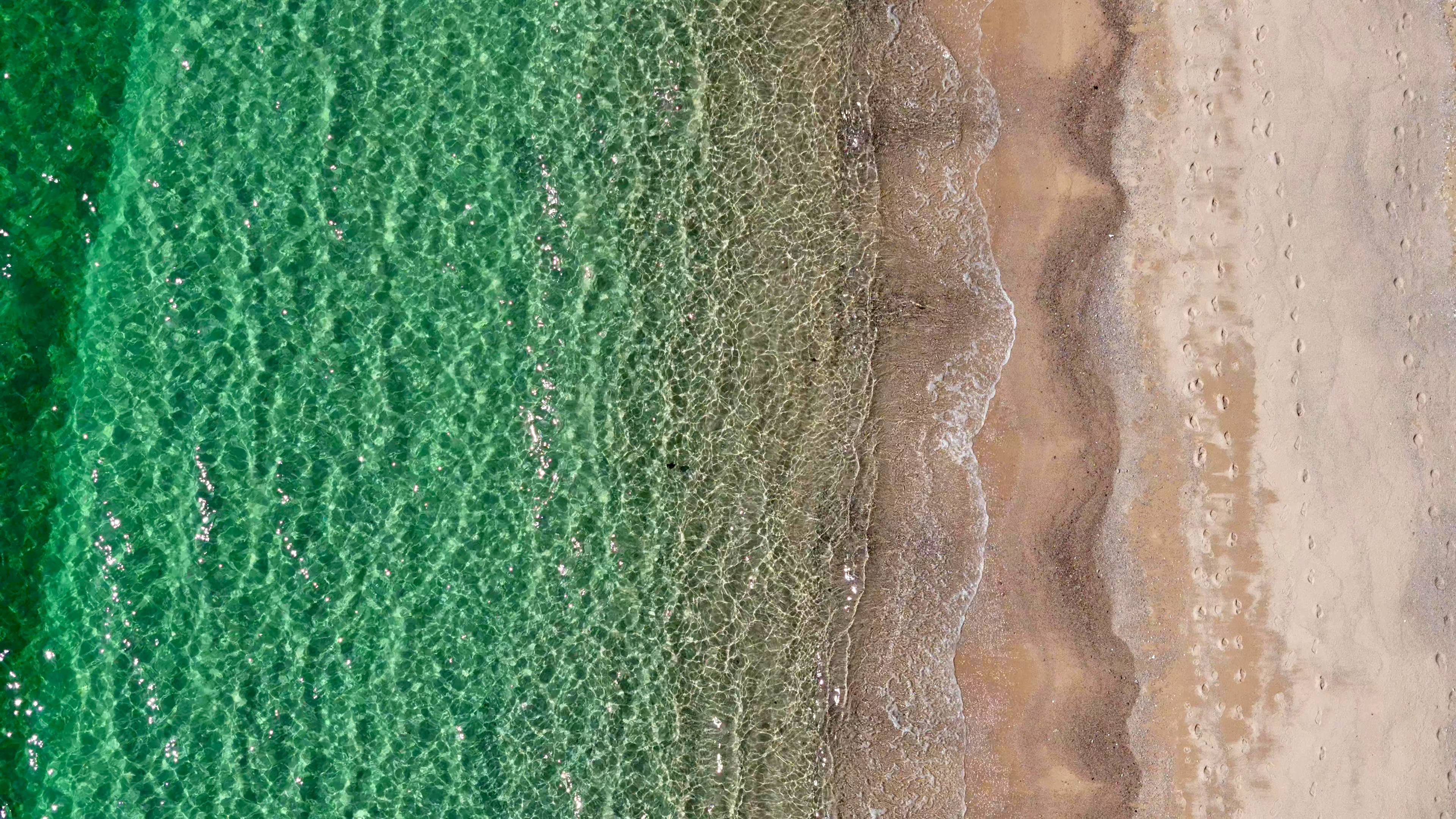 Straight down on the shoreline, turquoise shallows meeting wet sand and a line of dark pebbles.