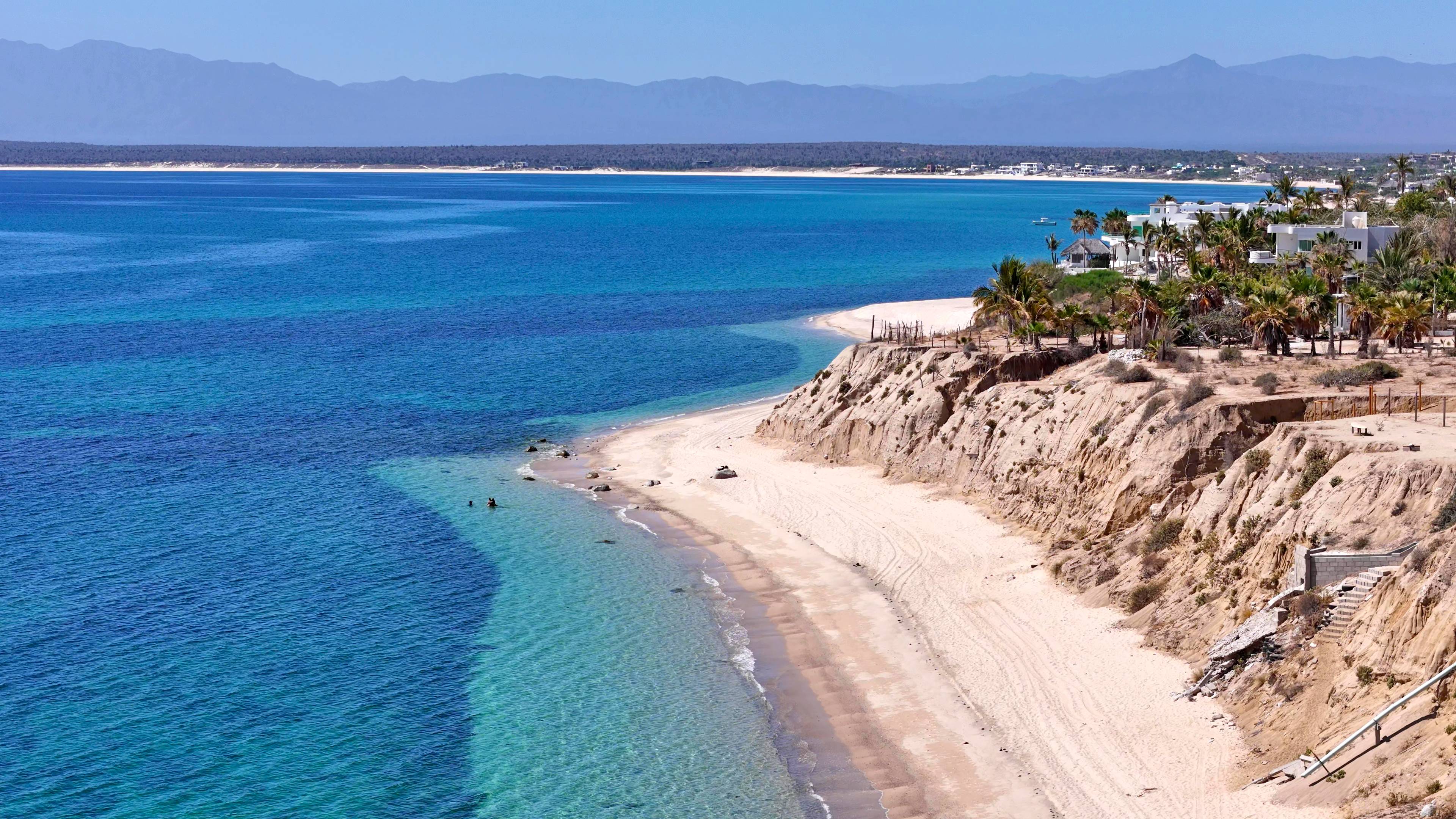 The bay from the air, turquoise shallows running along the sand below an eroded bluff.
